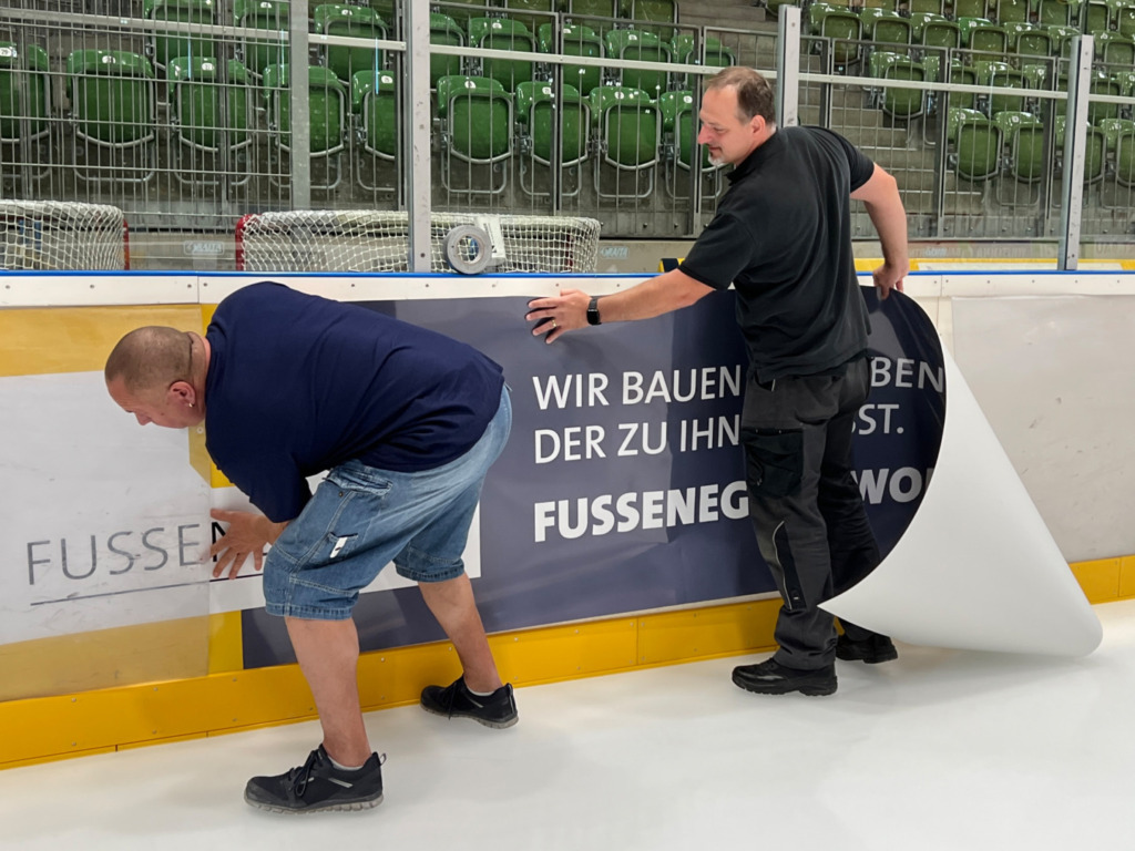 Mit Hilfe der beiden Eismeister zog die Fussenegger Wohnbau GmbH über die neue Bandenwerbung in die CHG Arena ein. Bild: Fussenegger Wohnbau GmbH